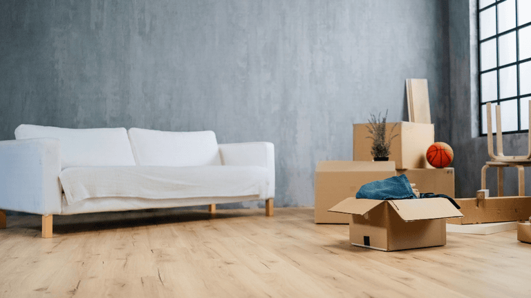 This image depicts a minimalist, partially furnished room with a white couch covered in a light cloth. The space appears to be in the midst of a move or unpacking process. Several cardboard boxes are placed around the room, some of them opened, with items like clothing, a basketball, and a plant visible. There is also a wooden chair and a pair of shoes. The large window and natural wood floor add a touch of warmth to the otherwise empty space.