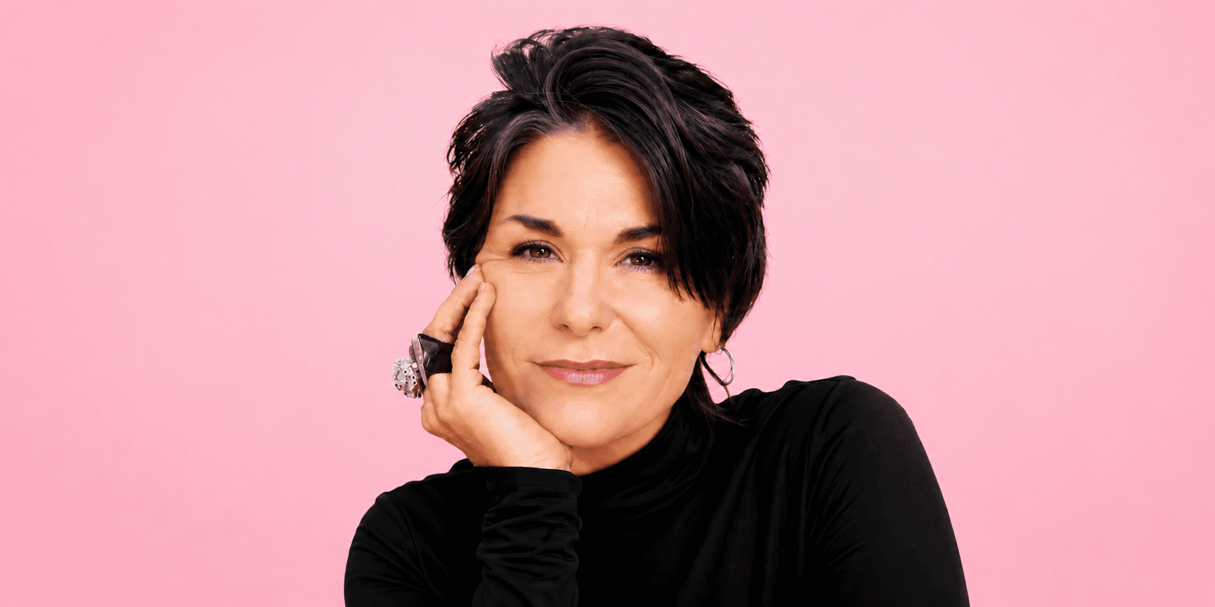 Woman with short dark hair wearing a black turtleneck poses against a pink background