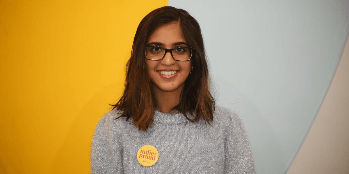 A young woman with glasses wearing a fuzzy sweater and a badge smiles at the camera