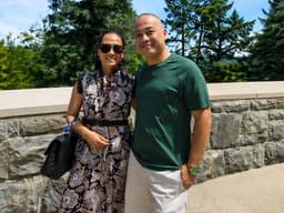 Logan Klink and his wife smiling together outdoors near a stone wall and trees.