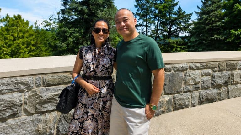 Logan Klink and his wife smiling together outdoors near a stone wall and trees.
