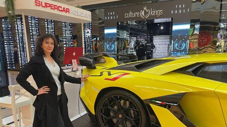 A woman stands confidently beside a bright yellow sports car in a mall, near a booth labeled "Supercar Holidays." The atmosphere is sleek and modern.