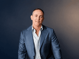 TJ Woodward wearing a blue blazer during a professional portrait against a dark background.