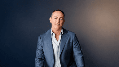 TJ Woodward wearing a blue blazer during a professional portrait against a dark background.