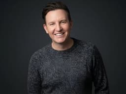 Zach Randles-Friedman smiling in a dark gray sweater against a dark studio background.