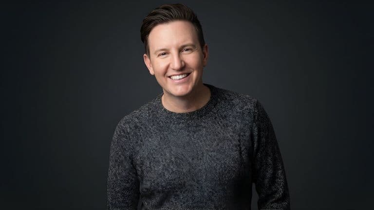 Zach Randles-Friedman smiling in a dark gray sweater against a dark studio background.