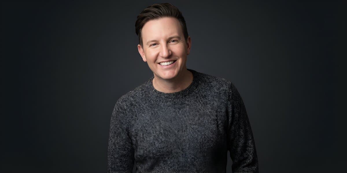 Zach Randles-Friedman smiling in a dark gray sweater against a dark studio background.