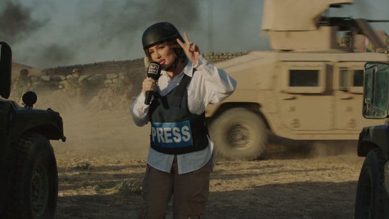A journalist wearing a helmet and press vest reports from a conflict zone, with military vehicles in the background.
