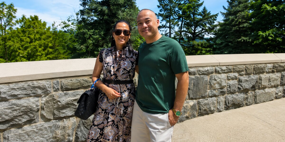 Logan Klink and his wife smiling together outdoors near a stone wall and trees.