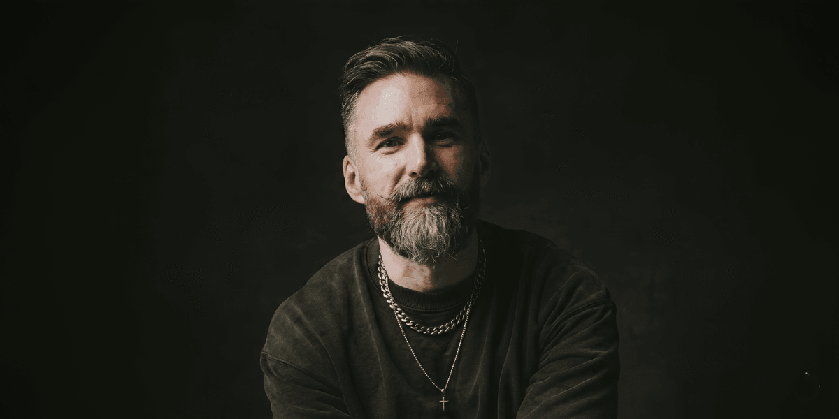 Portrait of Matt Jacob in a dark studio, wearing layered necklaces, smiling toward the camera.