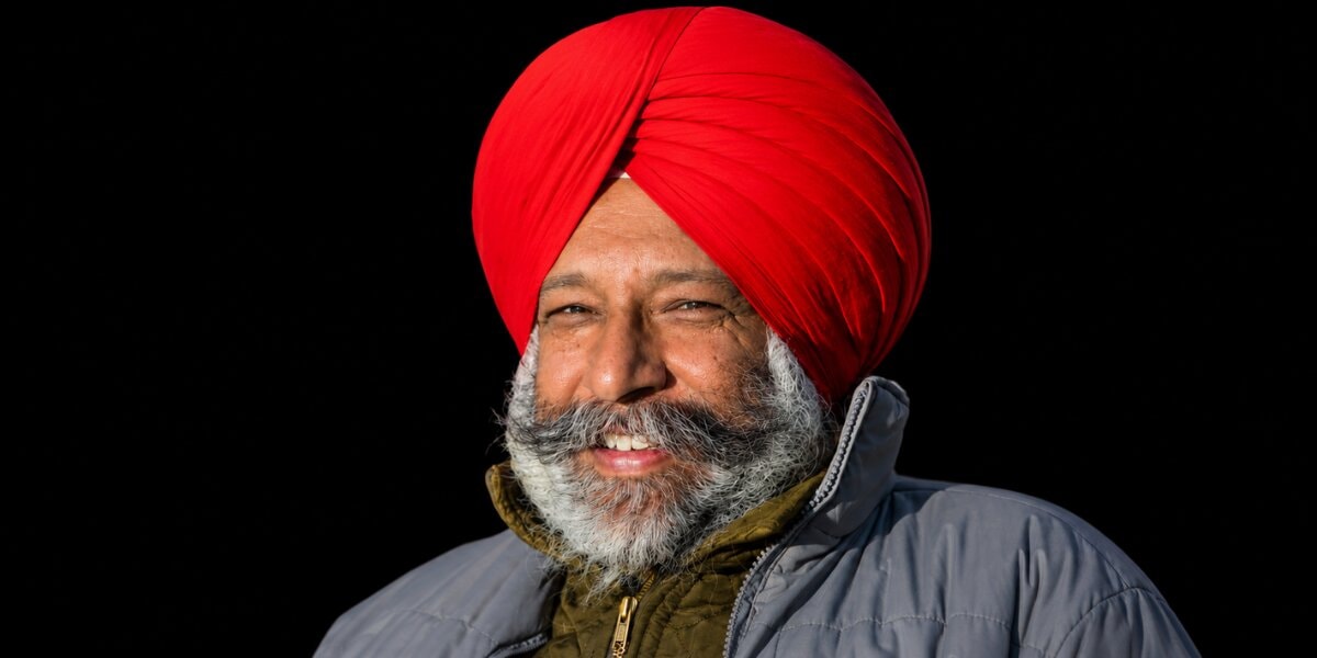 Portrait of smiling Sikh man in red turban and winter jacket against black background.