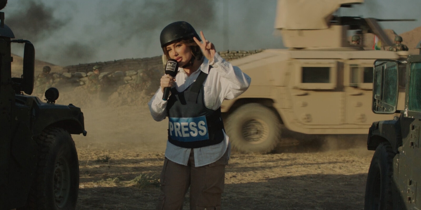 A journalist wearing a helmet and press vest reports from a conflict zone, with military vehicles in the background.