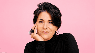 Woman with short dark hair wearing a black turtleneck poses against a pink background