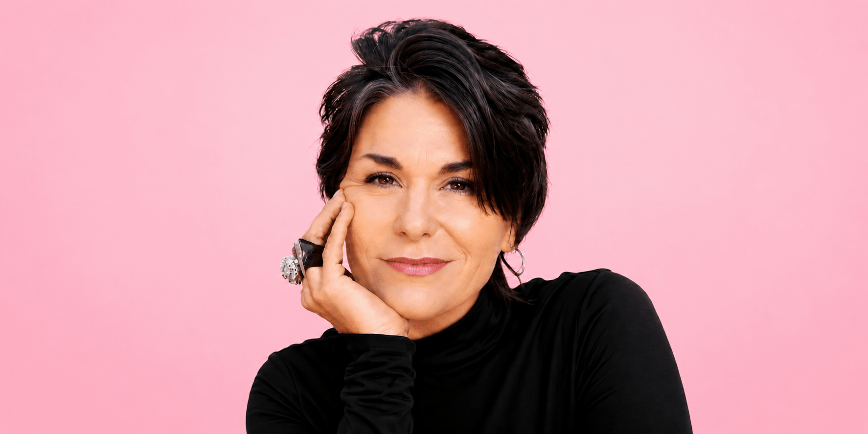 Woman with short dark hair wearing a black turtleneck poses against a pink background