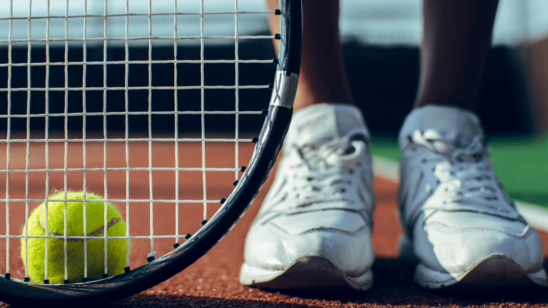 Tennis racket and ball on court with player wearing white shoes in focus