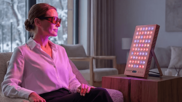 A woman with sunglasses sits in front of a red light therapy device.