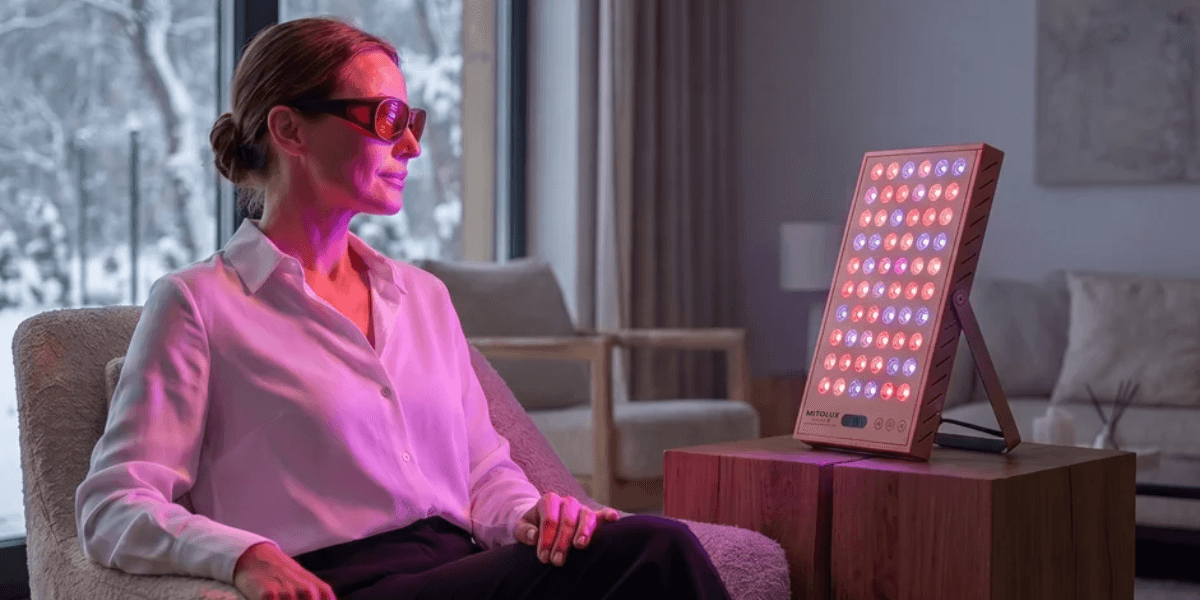 A woman with sunglasses sits in front of a red light therapy device.