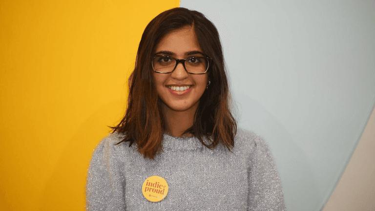 A young woman with glasses wearing a fuzzy sweater and a badge smiles at the camera