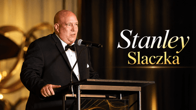 Man in a tuxedo speaking at a podium during an event with golden decor