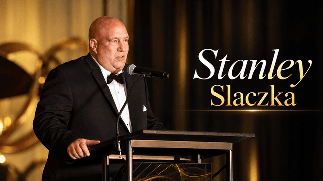 Man in a tuxedo speaking at a podium during an event with golden decor