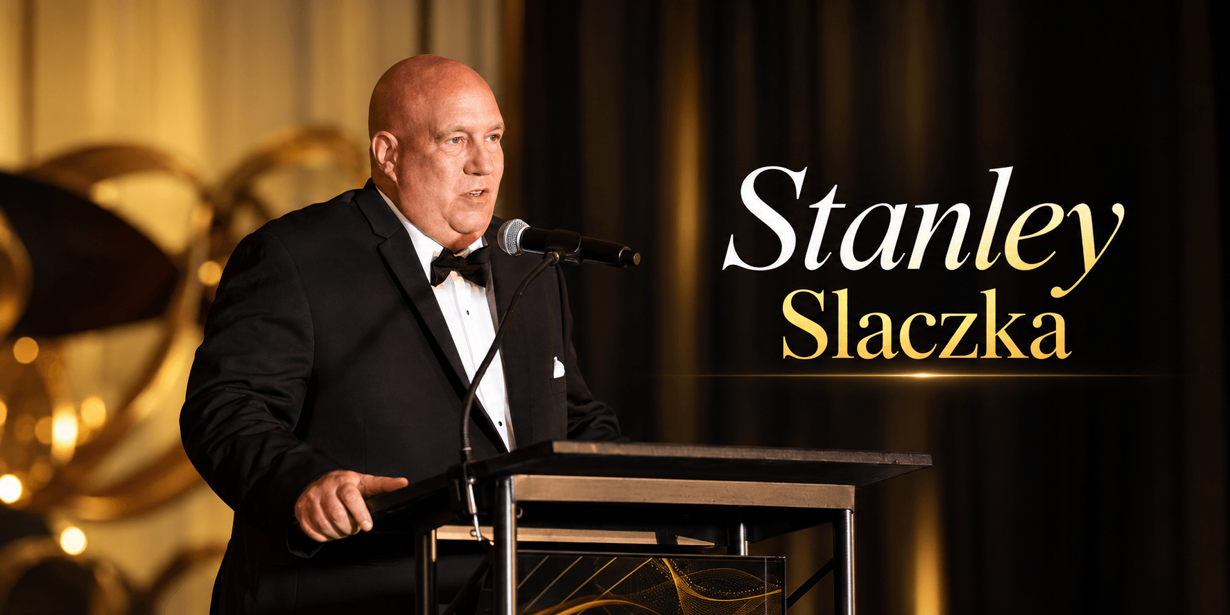 Man in a tuxedo speaking at a podium during an event with golden decor