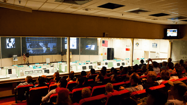 Interior of a space control room with audience and displays, simulating a historic mission