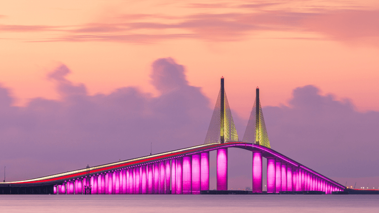 Vibrant bridge illuminated in pink against a pastel sunset sky over calm waters