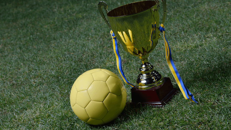 Yellow soccer ball beside a shiny trophy with blue and yellow ribbons on grass