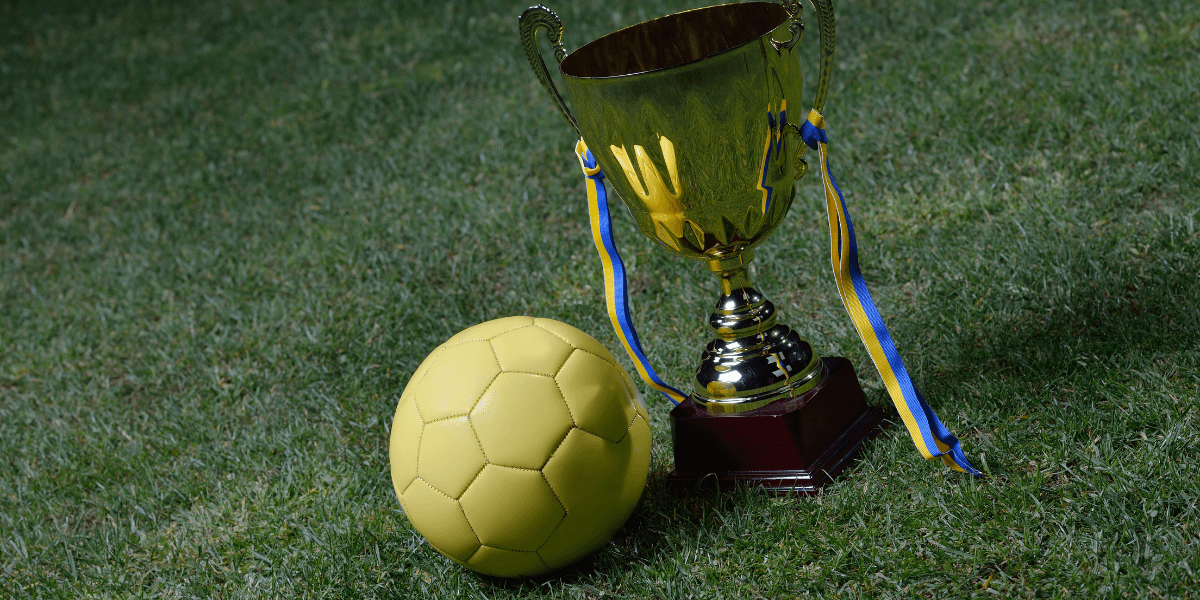 Yellow soccer ball beside a shiny trophy with blue and yellow ribbons on grass
