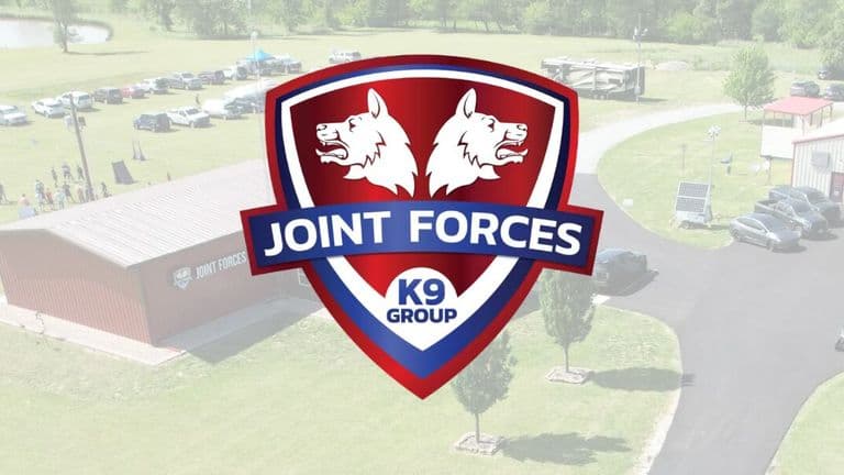 Shield-shaped logo with two white wolf heads on a red background, overlayed on an aerial view of a grassy area with buildings, parked cars, and trees. The words "Joint Forces K9 Group" are prominent.