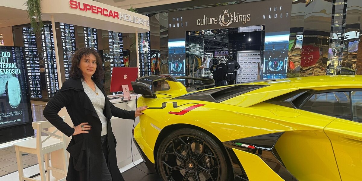 A woman stands confidently beside a bright yellow sports car in a mall, near a booth labeled "Supercar Holidays." The atmosphere is sleek and modern.