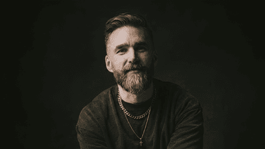 Portrait of Matt Jacob in a dark studio, wearing layered necklaces, smiling toward the camera.