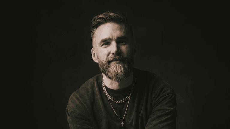Portrait of Matt Jacob in a dark studio, wearing layered necklaces, smiling toward the camera.