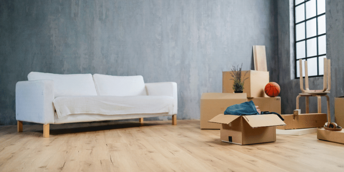 This image depicts a minimalist, partially furnished room with a white couch covered in a light cloth. The space appears to be in the midst of a move or unpacking process. Several cardboard boxes are placed around the room, some of them opened, with items like clothing, a basketball, and a plant visible. There is also a wooden chair and a pair of shoes. The large window and natural wood floor add a touch of warmth to the otherwise empty space.