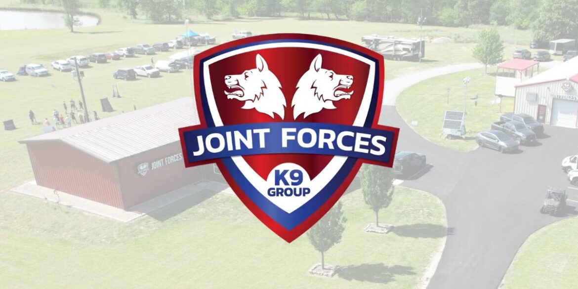 Shield-shaped logo with two white wolf heads on a red background, overlayed on an aerial view of a grassy area with buildings, parked cars, and trees. The words "Joint Forces K9 Group" are prominent.