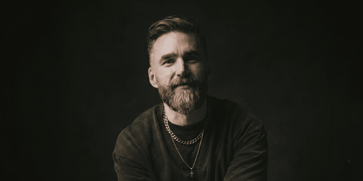 Portrait of Matt Jacob in a dark studio, wearing layered necklaces, smiling toward the camera.