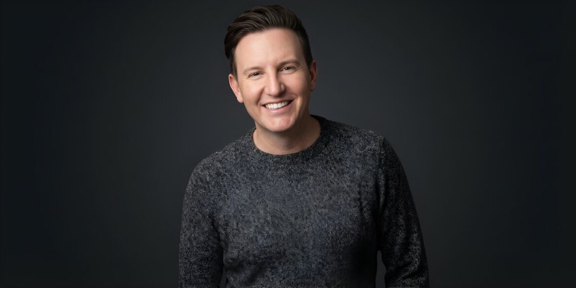 Zach Randles-Friedman smiling in a dark gray sweater against a dark studio background.