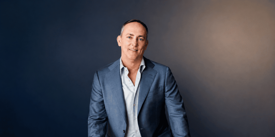 TJ Woodward wearing a blue blazer during a professional portrait against a dark background.